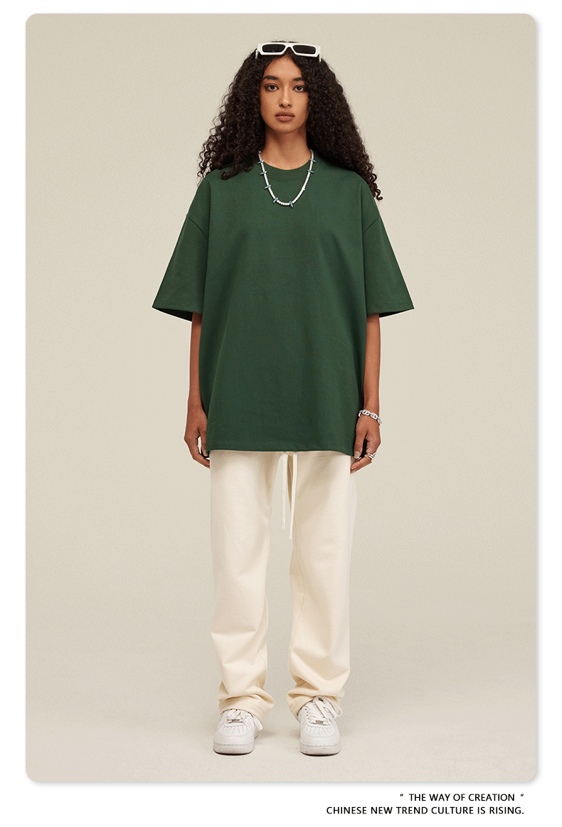 Woman wearing oversized dark green t-shirt with cream sweatpants, white sneakers and beaded necklace against neutral backdrop