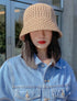 A woman wearing a khaki open-weave crochet bucket hat paired with a light blue denim jacket, standing outdoors near a modern cafe with a neutral background.