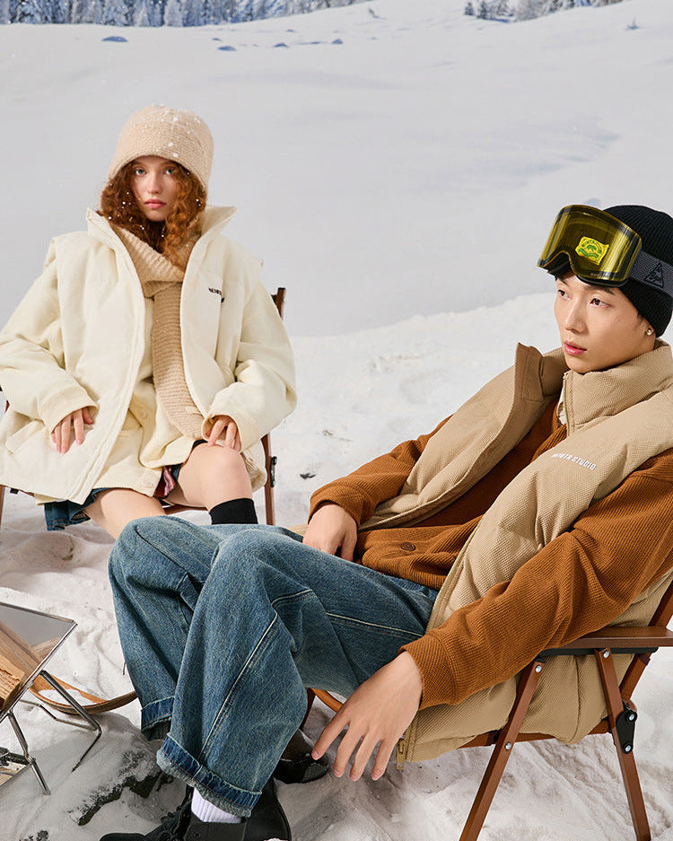 Models sitting in chairs on snowy mountain - woman in cream puffer and knit hat, man in tan puffer jacket with yellow ski goggles