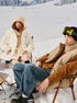 Models sitting in chairs on snowy mountain - woman in cream puffer and knit hat, man in tan puffer jacket with yellow ski goggles