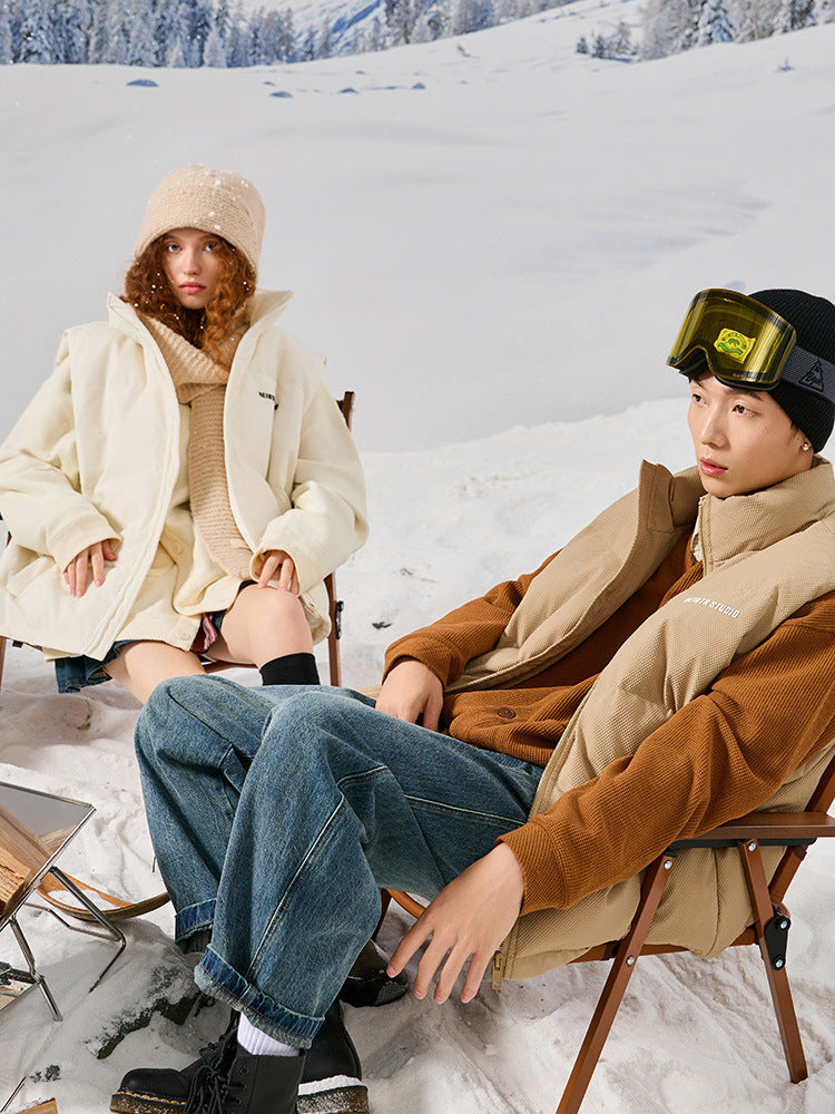 Models sitting in chairs on snowy mountain - woman in cream puffer and knit hat, man in tan puffer jacket with yellow ski goggles
