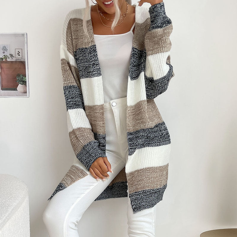 A woman smiling in colorblock knit cardigan with white, taupe and navy stripes paired with white tank top and jeans against minimal backdrop