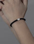A close-up shot of a black braided bracelet with a silver charm, worn on a person's wrist, against a muted background.