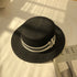 A black straw boater hat with a black-and-white ribbon band and silver charm, placed on a white sheet with sunlight casting shadows.