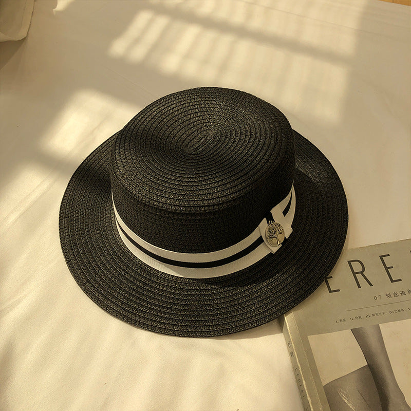 A black straw boater hat with a black-and-white ribbon band and silver charm, placed on a white sheet with sunlight casting shadows.