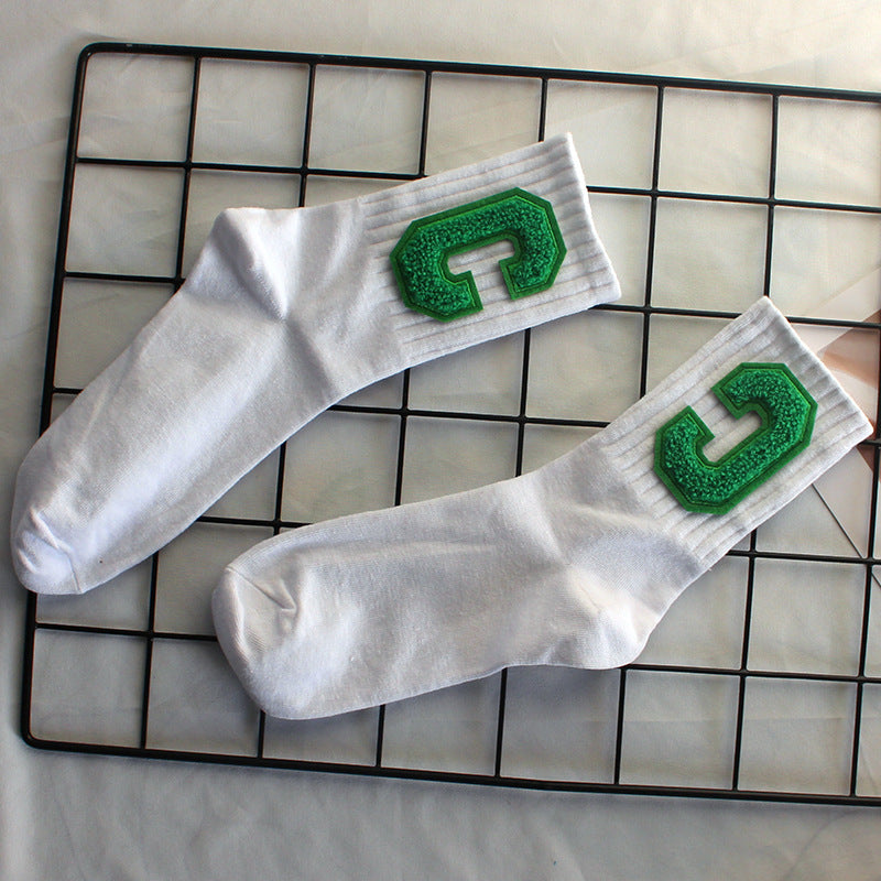 A pair of white varsity-style crew socks with green textured "C" patches is laid out on a black wire grid, positioned over a neutral fabric surface