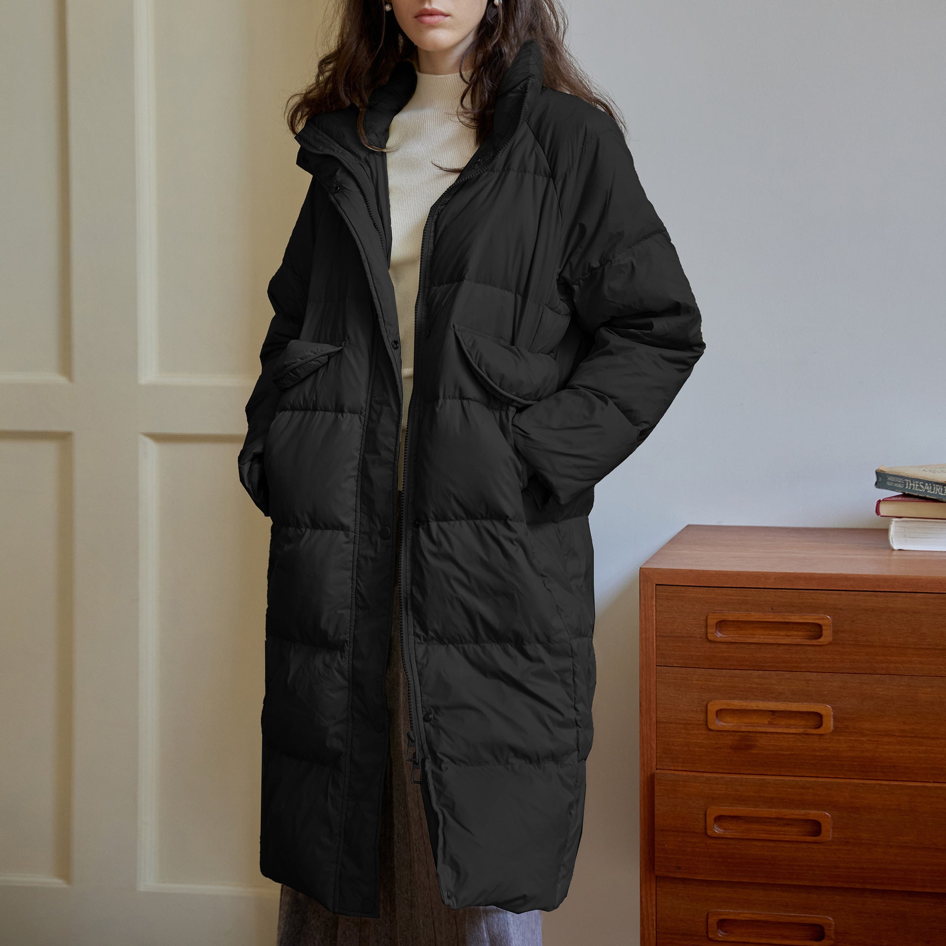 Black long puffer coat with flap pockets and high neck, worn with cream turtleneck against paneled wall