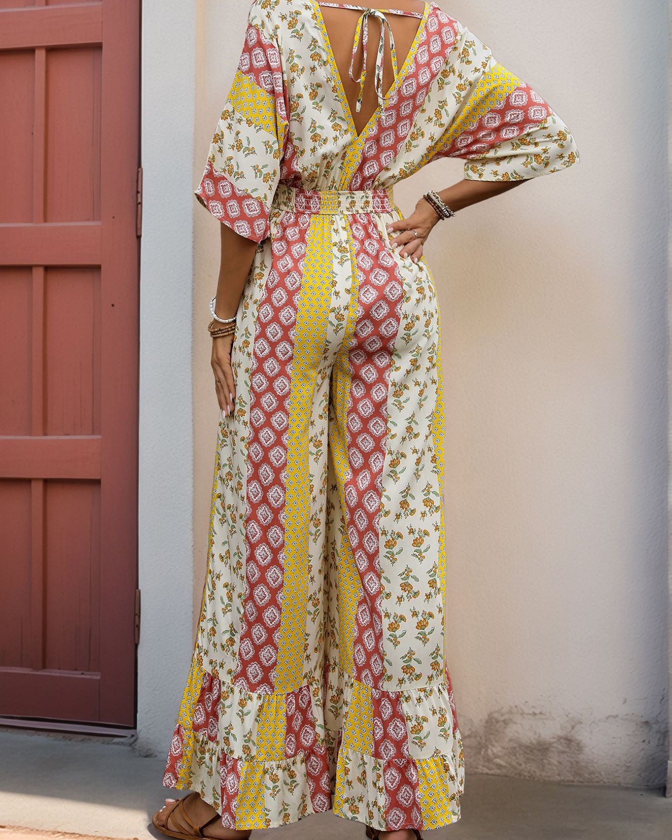 Back view of a woman in a V-back patchwork jumpsuit with a tie detail, loose wide-leg pants, and bohemian-inspired print.
