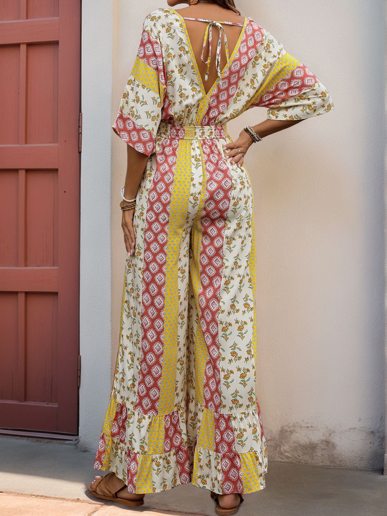 Back view of a woman in a V-back patchwork jumpsuit with a tie detail, loose wide-leg pants, and bohemian-inspired print.