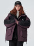 A woman with curly red hair wearing a Brooklyn cap clasps hands over her plum and black All Drifting jacket against white background