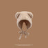 A beige teddy bear bonnet with rounded ears and tie closure photographed against brown background, featuring soft plush texture
