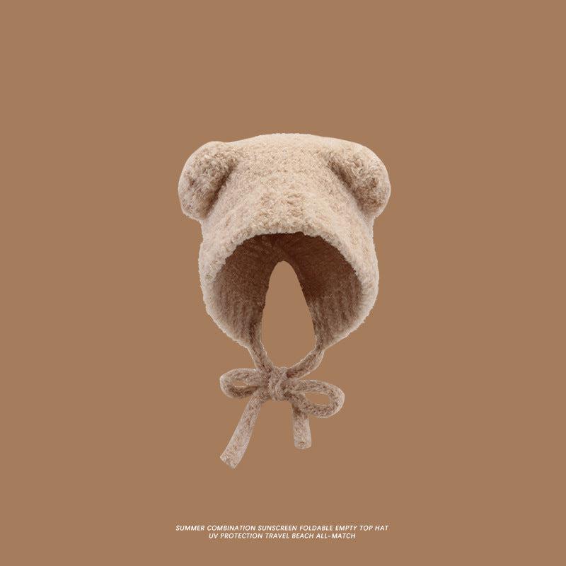 A beige teddy bear bonnet with rounded ears and tie closure photographed against brown background, featuring soft plush texture
