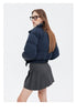 A woman stands with her back turned, wearing a navy puffer jacket with a plaid collar, a gray pleated skirt, and brown platform boots