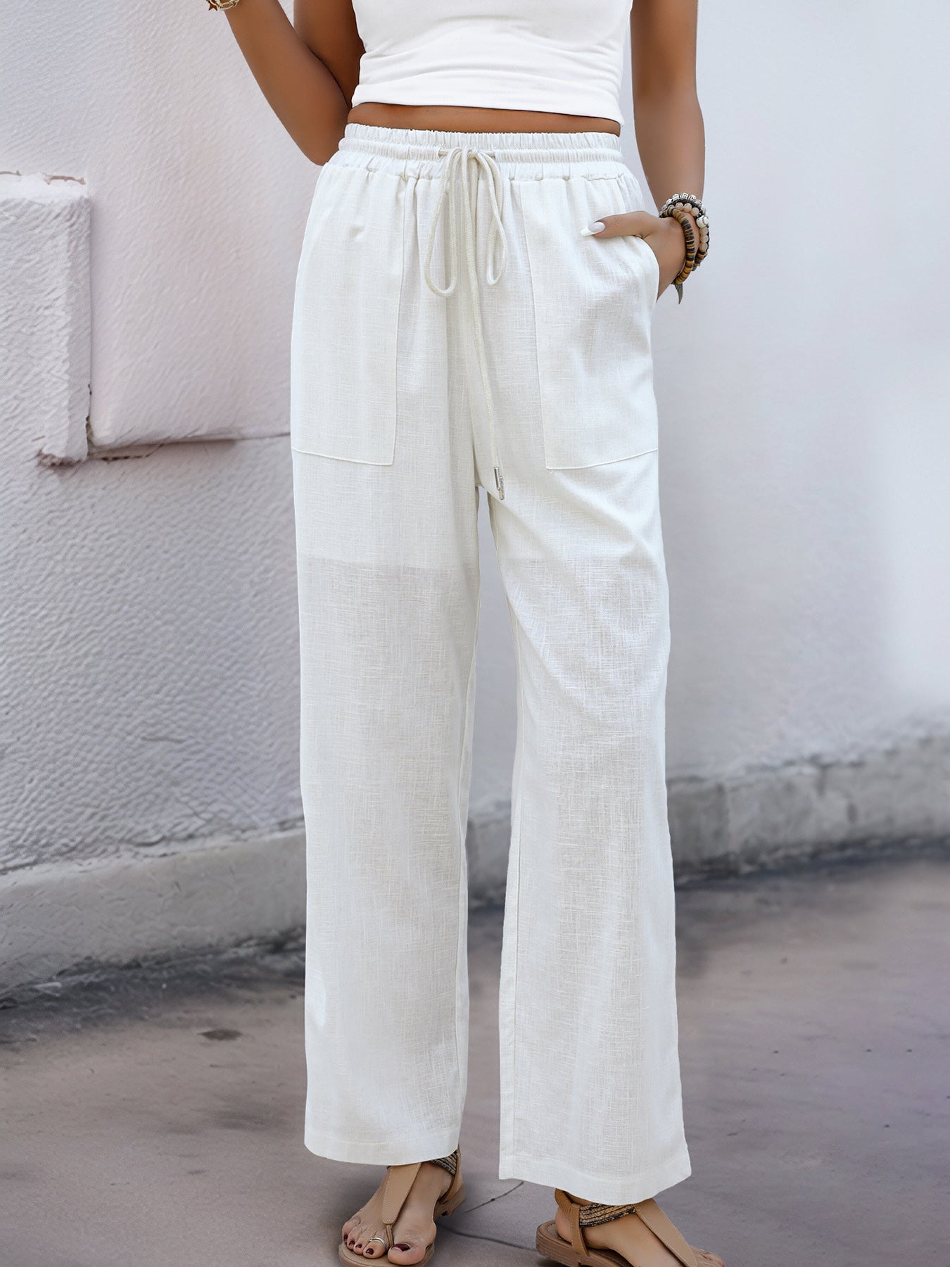 Woman standing with one hand in the pocket of her white wide-leg linen pants, styled with a white tank and bracelets.