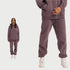 Model in purple hoodie sweatsuit with matching joggers, white sneakers, and gold necklace against white background