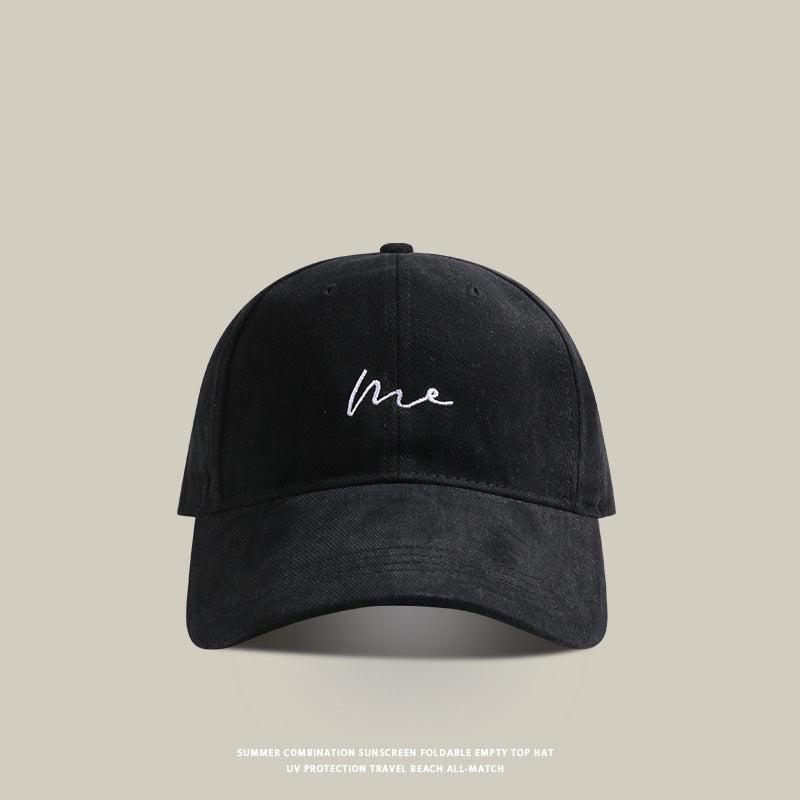 Black baseball cap with 'Me' embroidered in white cursive, featuring a curved brim and adjustable strap