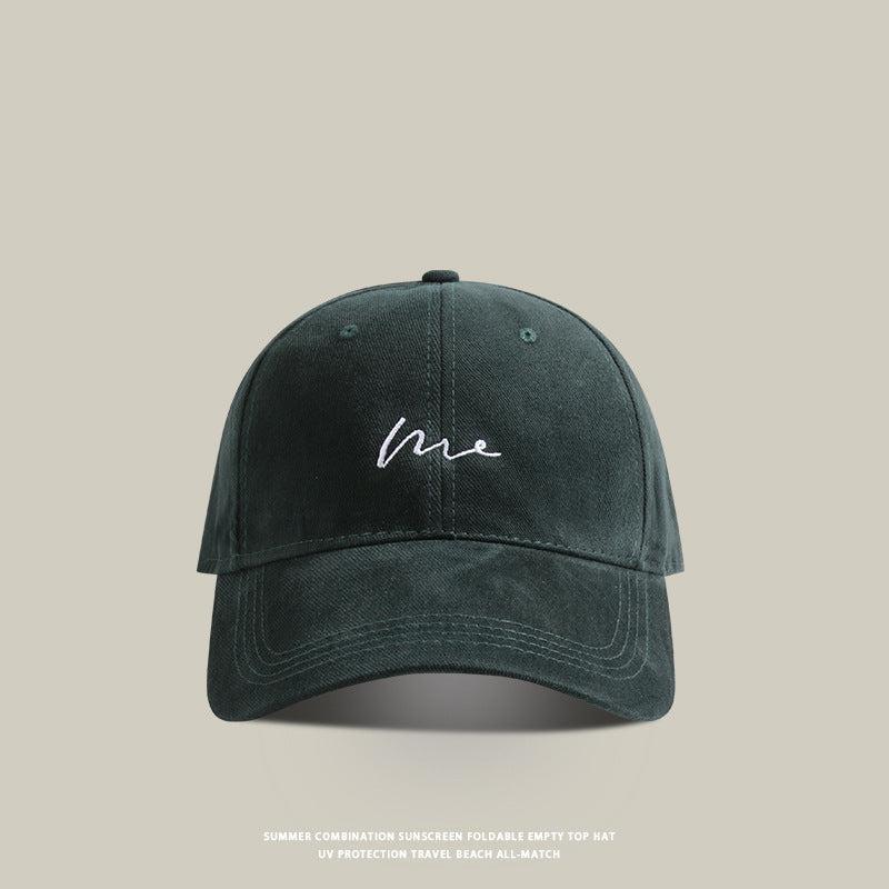 Dark green baseball cap with 'Me' embroidered in white cursive, classic curved brim, and soft washed fabric