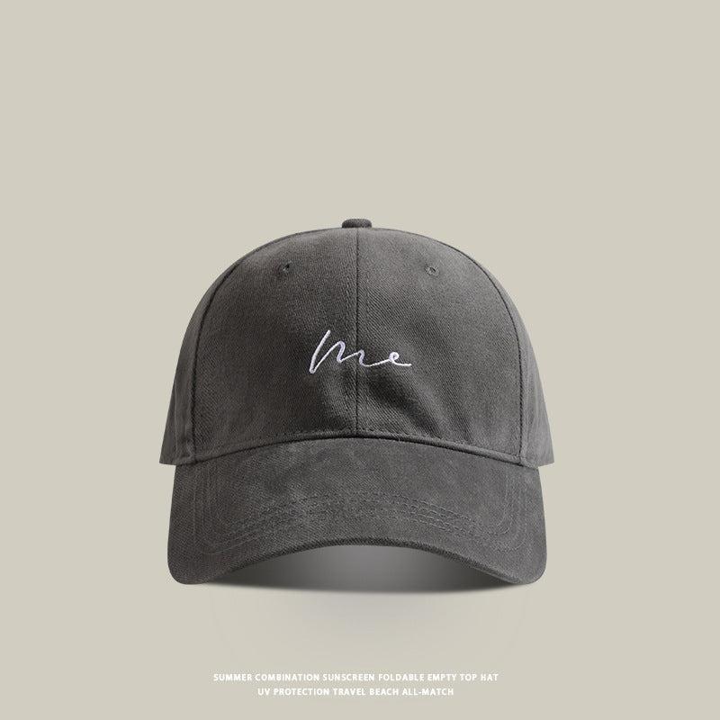 Gray baseball cap with 'Me' embroidered in white cursive, featuring a curved brim and soft cotton material
