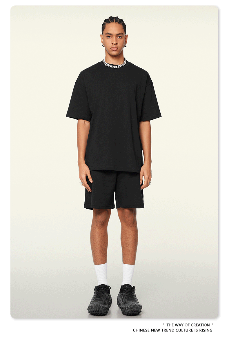 A man with braided hair wearing a black oversized t-shirt and shorts set, styled with silver chain necklace, white socks, and black sneaker
