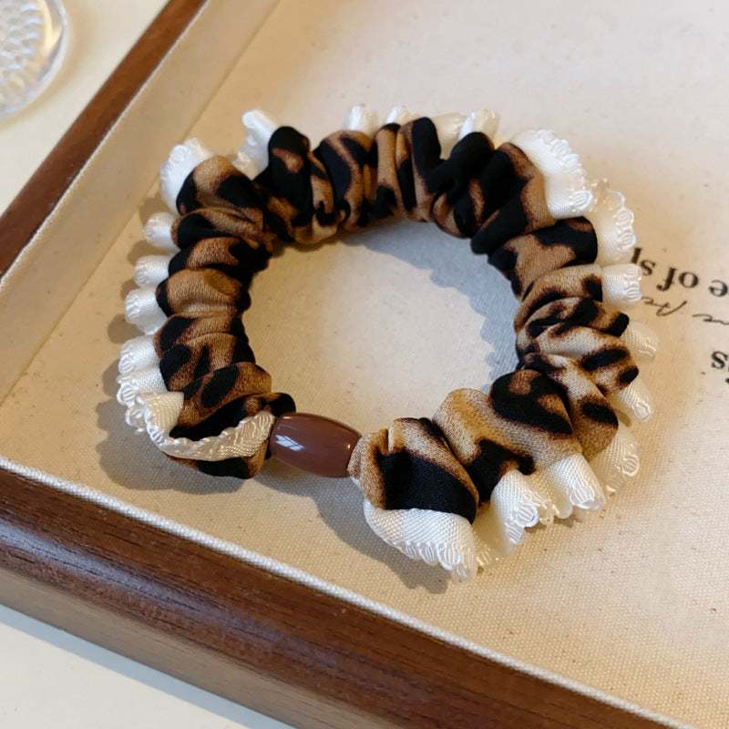 Detailed view of leopard print scrunchie with white ruffle trim and brown bead accent on display tray against neutral background