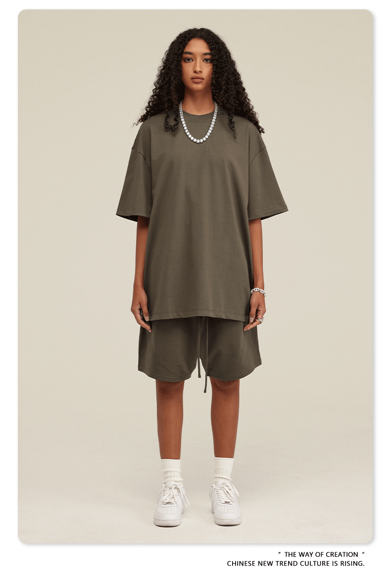 A woman with curly hair wearing an olive green oversized t-shirt and matching shorts set, styled with white socks, sneakers, and a pearl necklace