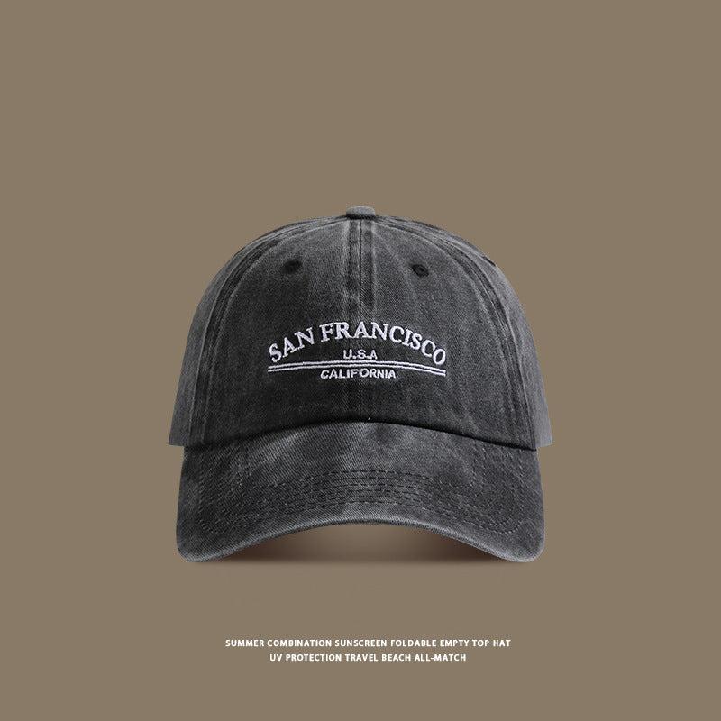 A dark gray washed cotton baseball cap with white 'San Francisco USA California' embroidery displayed against taupe background with travel text