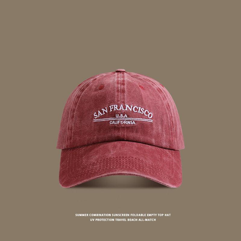 A brick red washed cotton baseball cap with white 'San Francisco USA California' embroidery displayed against taupe background with beach text