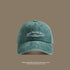 A sage green washed cotton baseball cap with white 'San Francisco USA California' embroidery displayed against taupe background with travel text