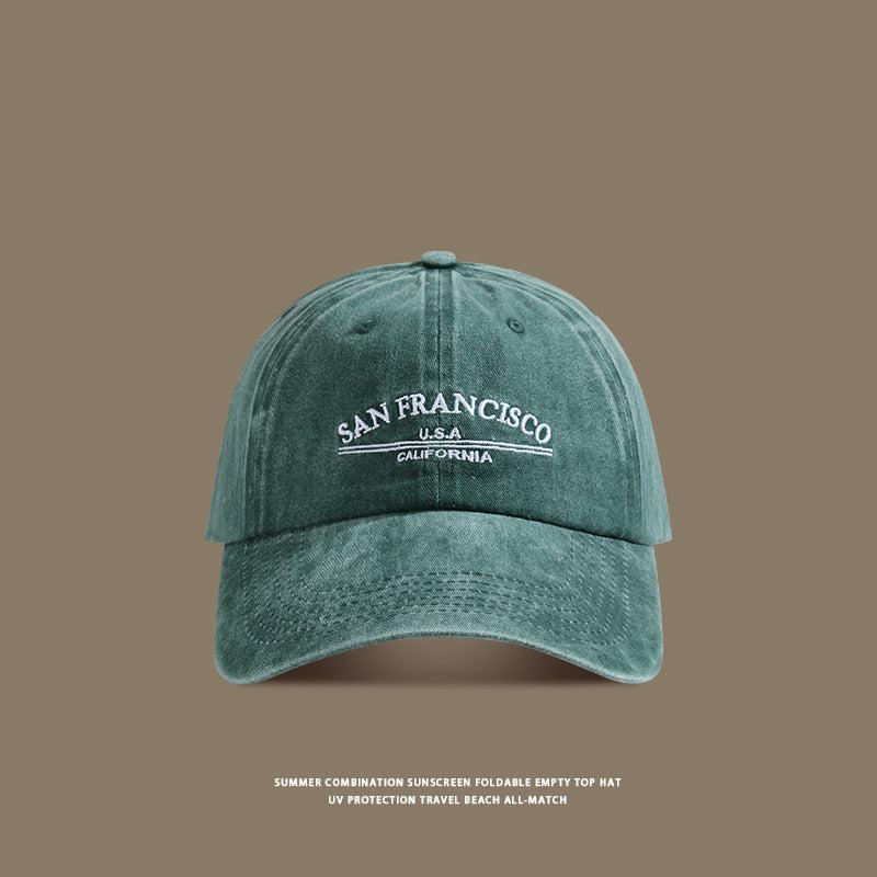 A sage green washed cotton baseball cap with white 'San Francisco USA California' embroidery displayed against taupe background with travel text