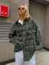 Forest green windowpane plaid shacket with white pants and oval sunglasses, urban casual style against industrial backdrop