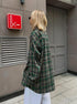 Side view of forest green plaid oversized shirt with white pants, showing relaxed silhouette against industrial setting