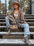 A woman is sitting on outdoor metal stairs wearing a brown plaid oversized shirt, white strappy crop top, blue jeans, cowboy boots, and a beige wide-brim hat, holding a tan handbag