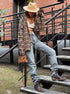 A woman is wearing a brown plaid oversized shirt, white strappy crop top, blue jeans, cowboy boots, and a beige wide-brim hat, standing on a metal staircase