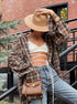 A woman is adjusting a beige wide-brim hat while wearing a brown plaid oversized shirt, white strappy crop top, and blue jeans, posing on an outdoor staircase