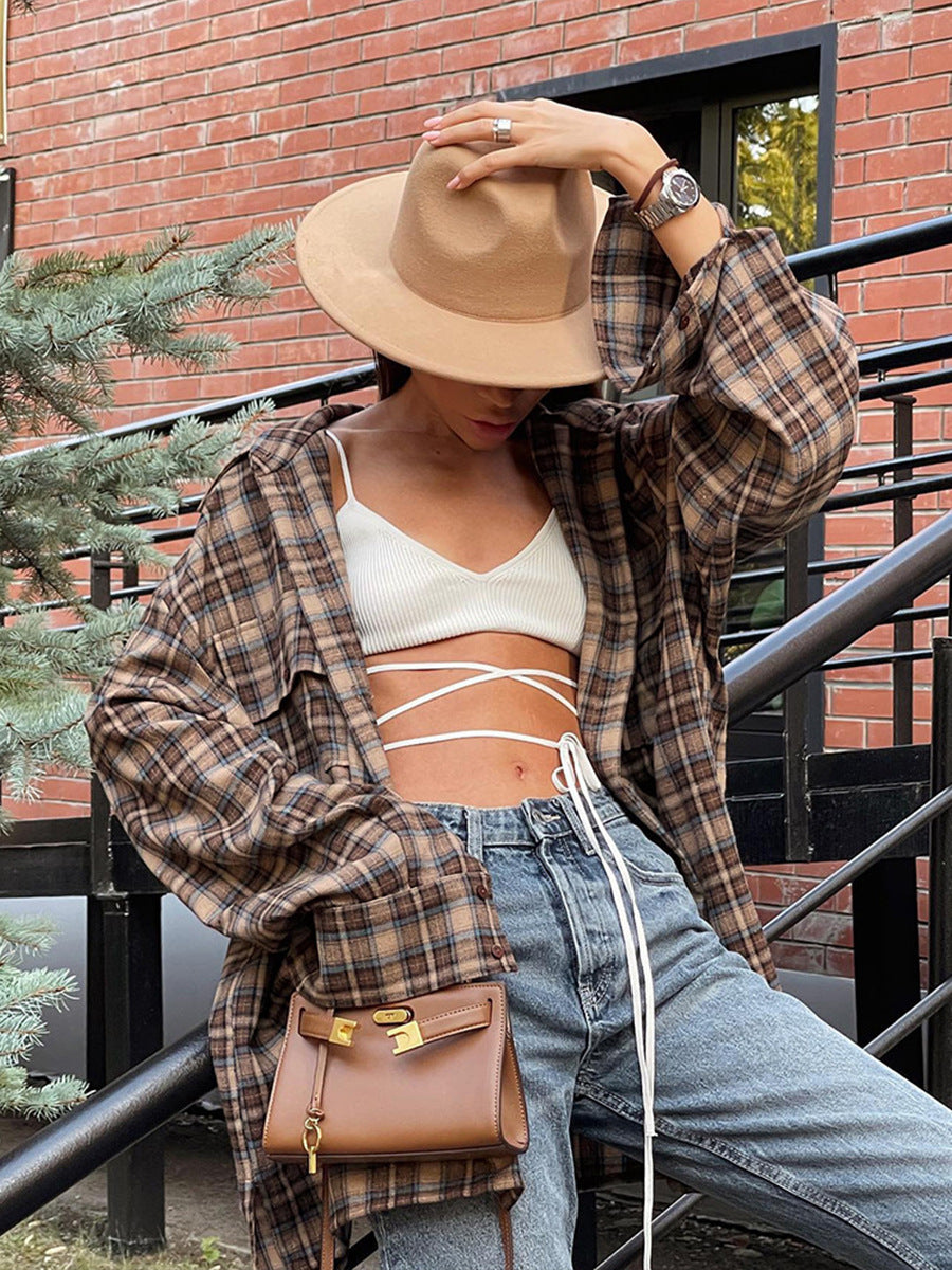 A woman is adjusting a beige wide-brim hat while wearing a brown plaid oversized shirt, white strappy crop top, and blue jeans, posing on an outdoor staircase