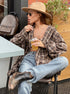 A woman is sitting at an outdoor café table wearing a brown plaid oversized shirt, a beige wide-brim hat, sunglasses, and cowboy boots, sipping a drink