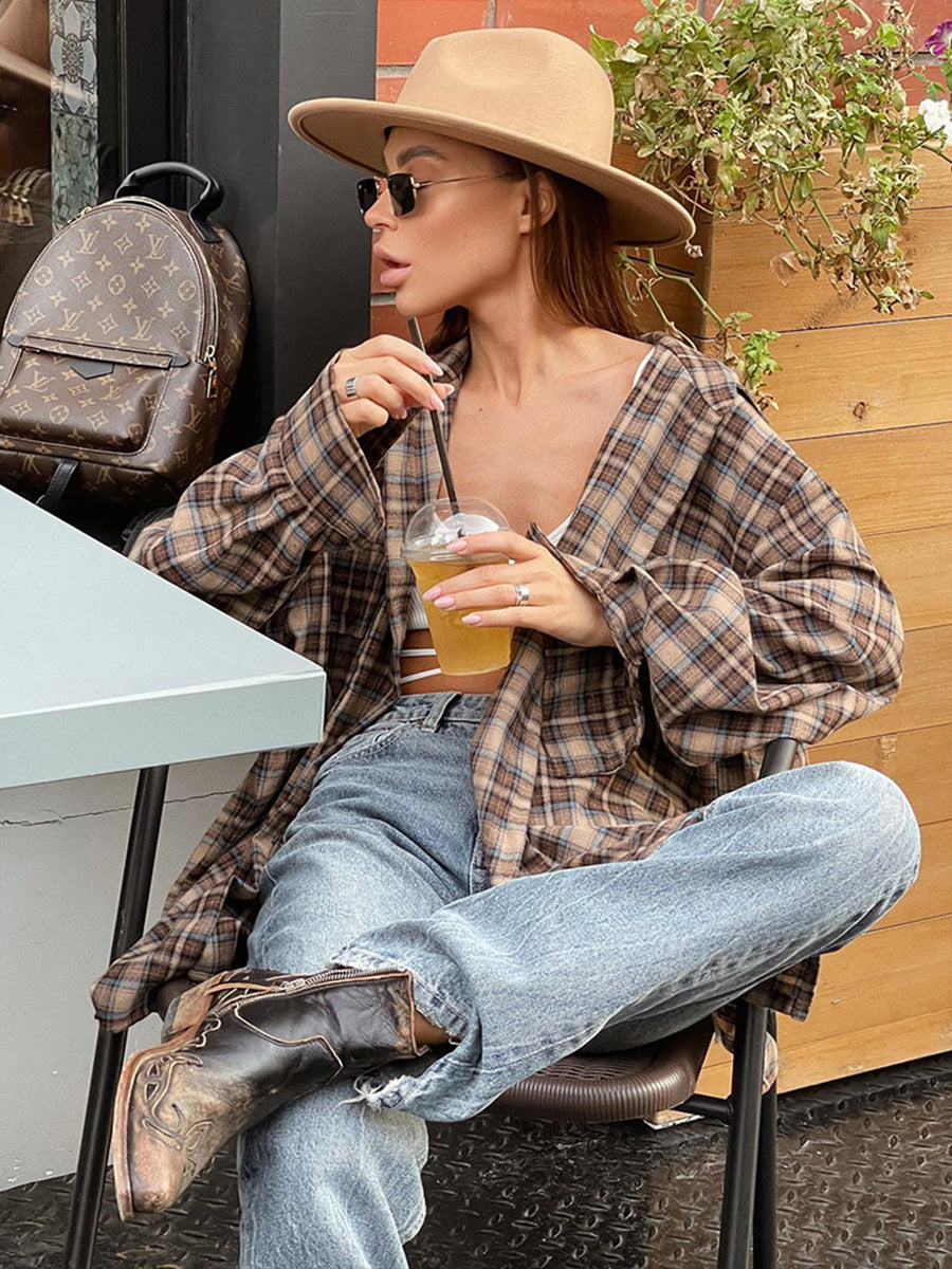 A woman is sitting at an outdoor café table wearing a brown plaid oversized shirt, a beige wide-brim hat, sunglasses, and cowboy boots, sipping a drink