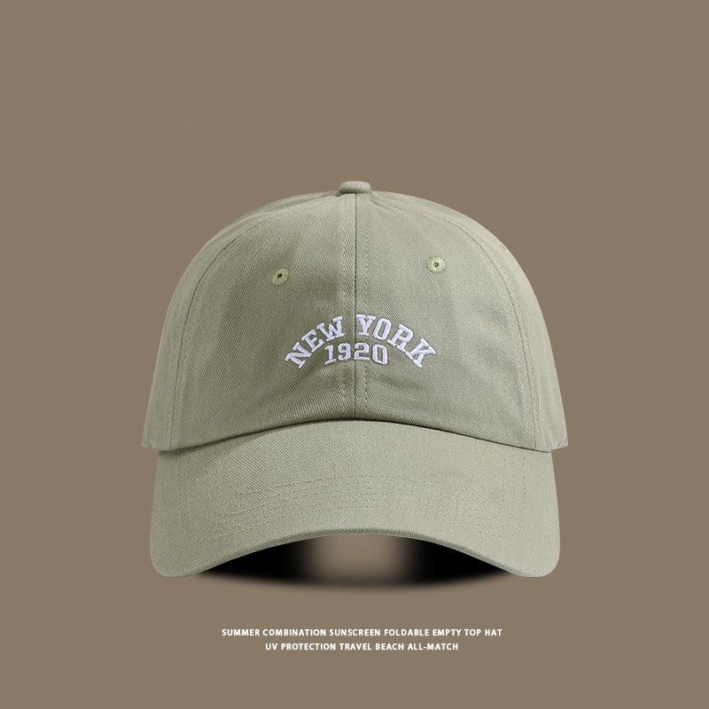 Olive green baseball cap featuring "New York 1920" embroidered in white, positioned against a neutral brown background