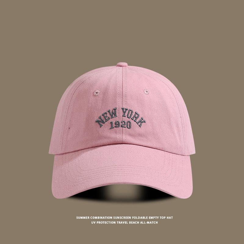 Soft pink baseball cap with "New York 1920" stitched in dark embroidery, set against a brown background