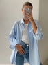 A woman is wearing a blue striped oversized button-up shirt over a white tank top with high-waisted jeans, posing with her phone