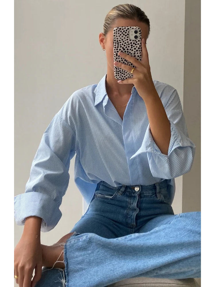 A woman is wearing a blue striped oversized button-up shirt with blue jeans, sitting cross-legged while taking a mirror selfie