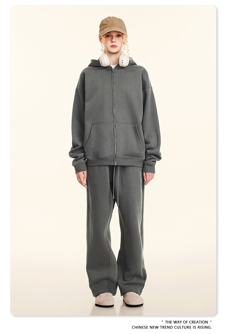 Blonde model wearing charcoal gray oversized zip-up hoodie and sweatpants with tan cap, white headphones and beige shoes against cream background