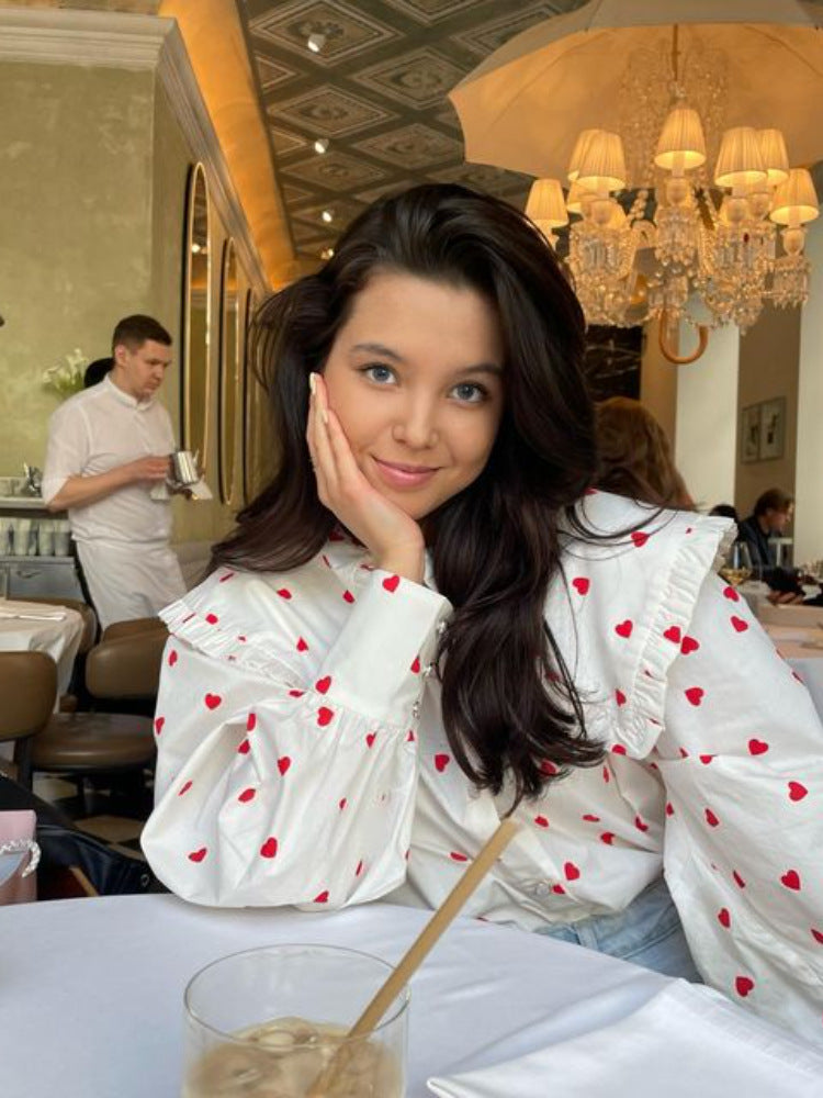 A woman wearing a white blouse with red hearts and ruffle collar sits at a restaurant table with drink, smiling with hand on face