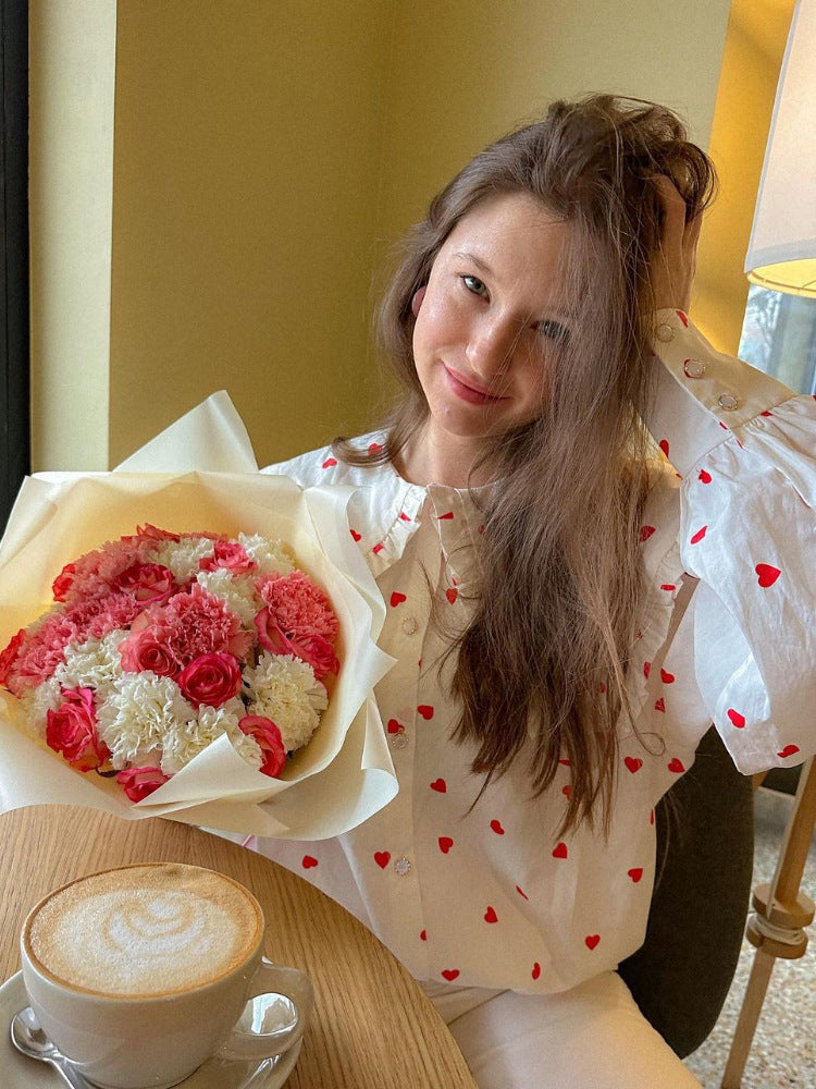 A woman wearing a white heart-pattern blouse with ruffled collar holds pink bouquet and sits at café table with latte in warm-toned setting