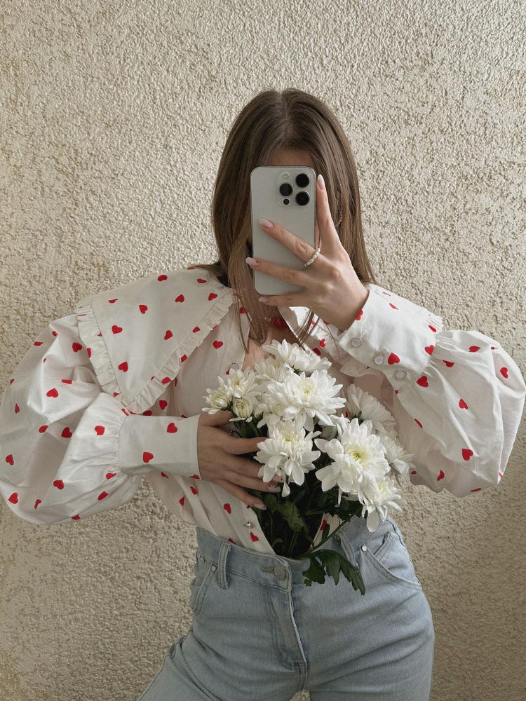 A woman taking mirror selfie wearing white heart-print blouse with ruffle collar and light denim jeans, holding white chrysanthemums