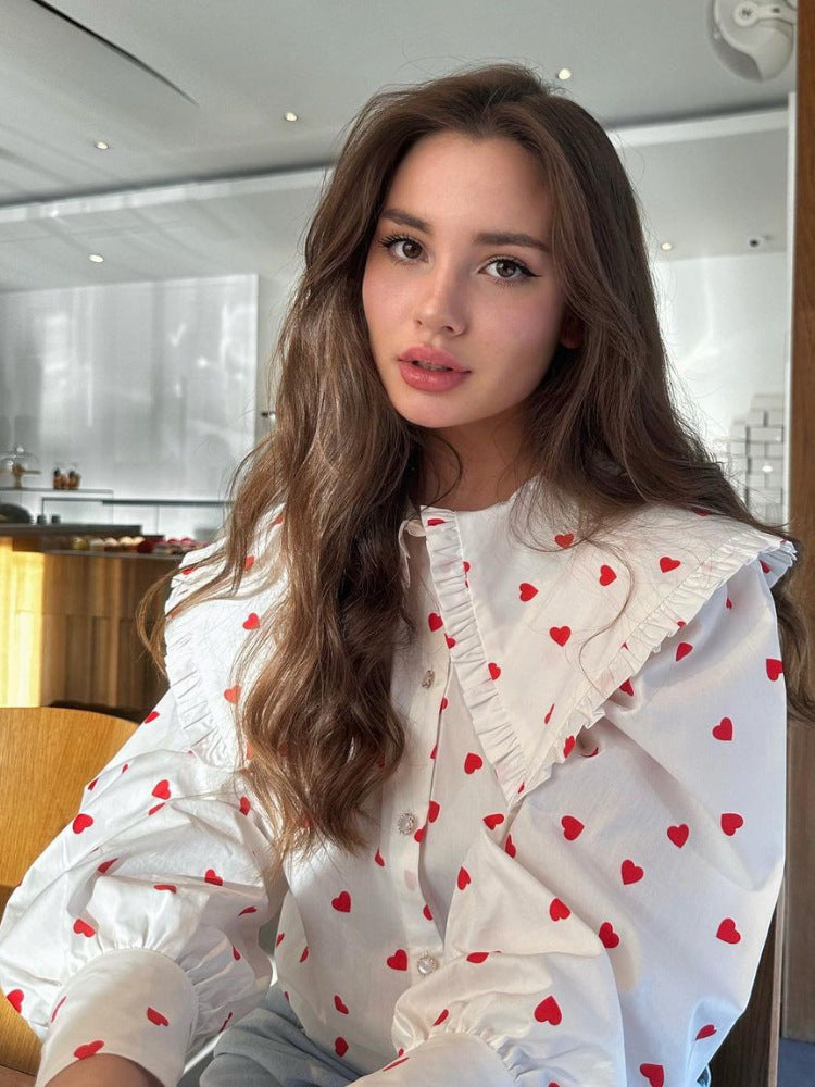 A woman wearing white red-heart print blouse with peter pan collar poses in bright modern interior with minimalist white background
