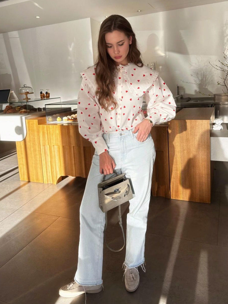 A woman wearing a white heart-print blouse with ruffle collar stands in a café, holding a gray handbag with light denim jeans and cream shoes
