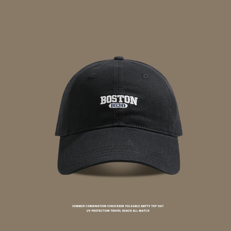 Black baseball cap with white Boston 1630 embroidered logo on taupe background