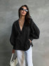 A woman is posing in an oversized black button-up shirt with balloon sleeves and white trousers, wearing rectangular sunglasses and delicate gold necklace