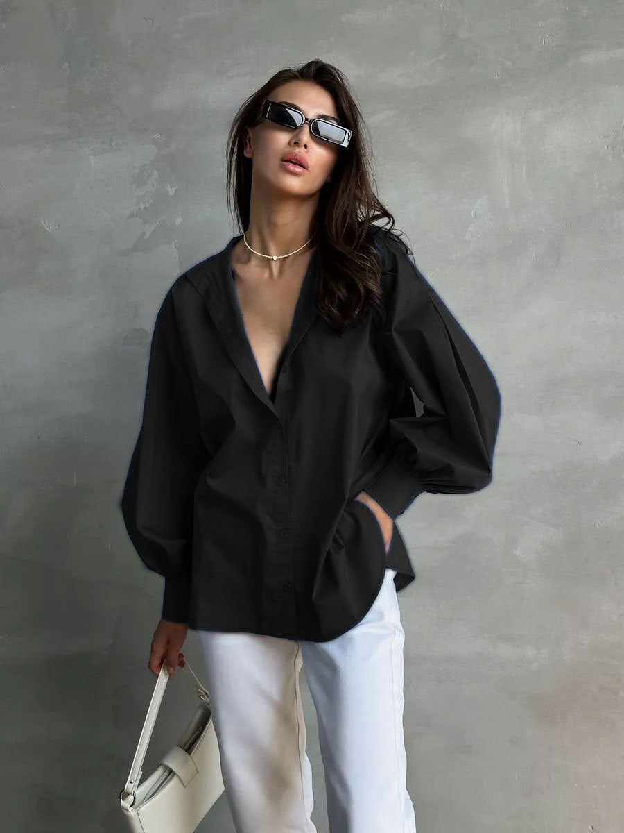 A woman is posing in an oversized black button-up shirt with balloon sleeves and white trousers, wearing rectangular sunglasses and delicate gold necklace